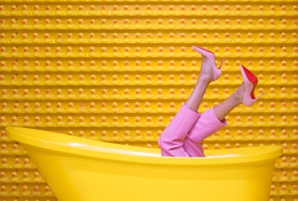Fargerikt bilde av gult badekar, gul vegg og to føtter opp av badekaret med rosa bukse og sko