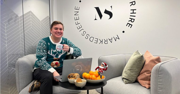 Markedsføreren Sindre ikledd julegenser sitter i stol og drikker en kaffe fra julekopp